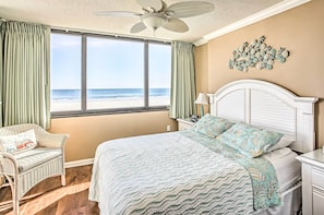 Une chambre avec une grande fenêtre donnant sur la plage, un lit recouvert d’une couvre-lit à motifs, une chaise en osier et un ventilateur de plafond.