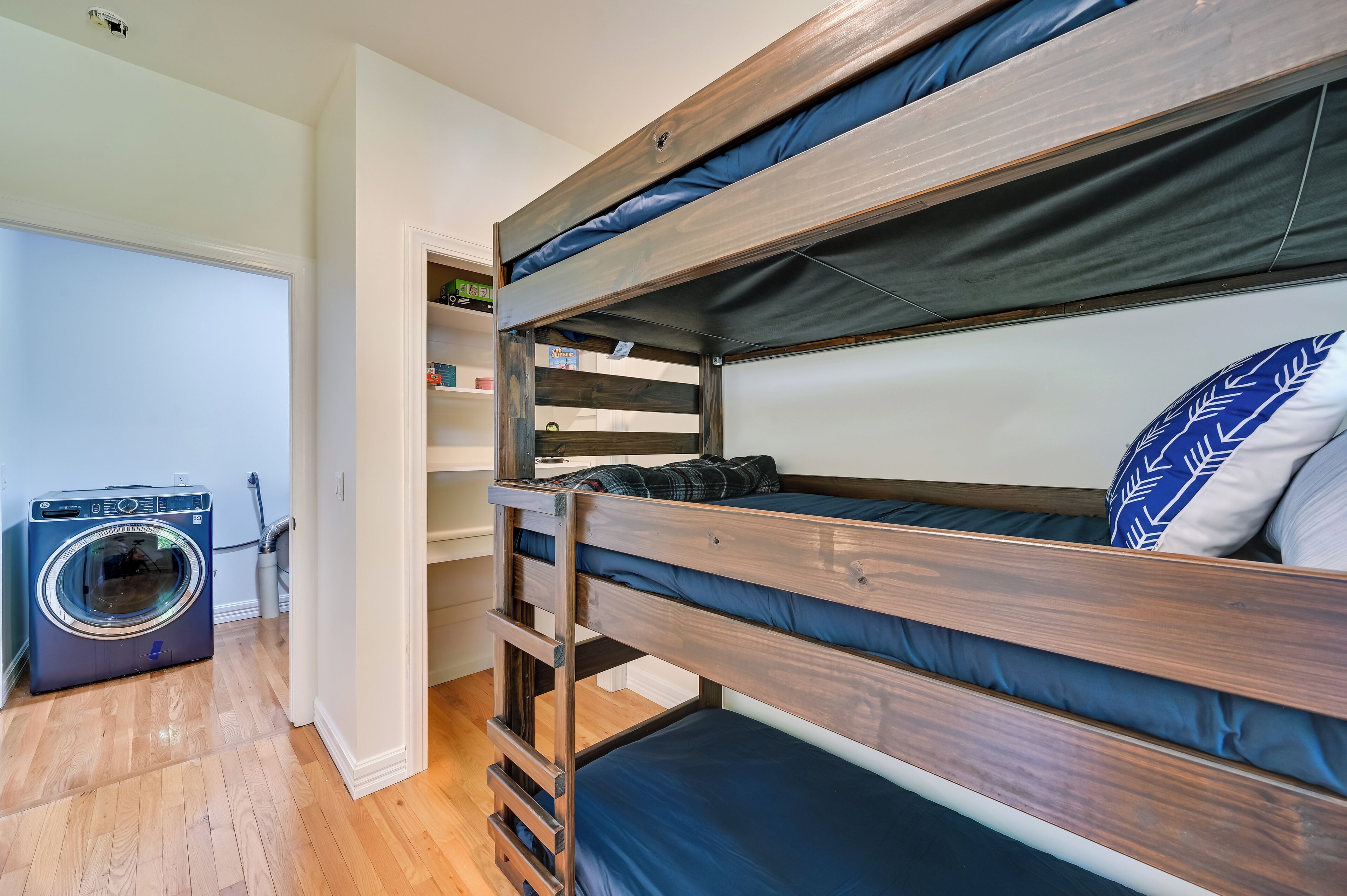 Een stapelbed met houten frame en blauw beddengoed, een wasmachine en een klein rek met diverse spullen.