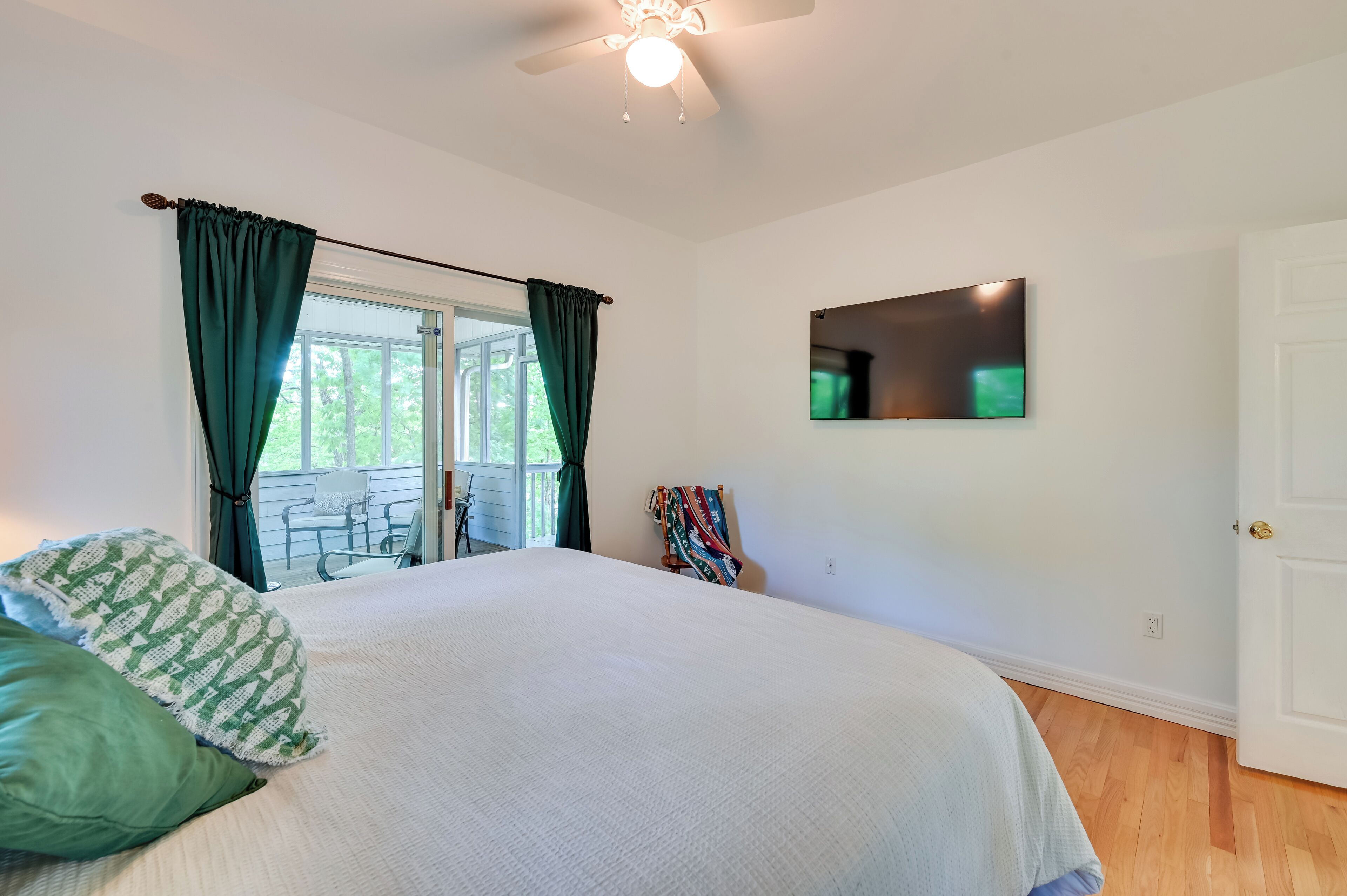 A bedroom with a bed, a ceiling fan, a window with curtains, a door, and a wall-mounted TV.