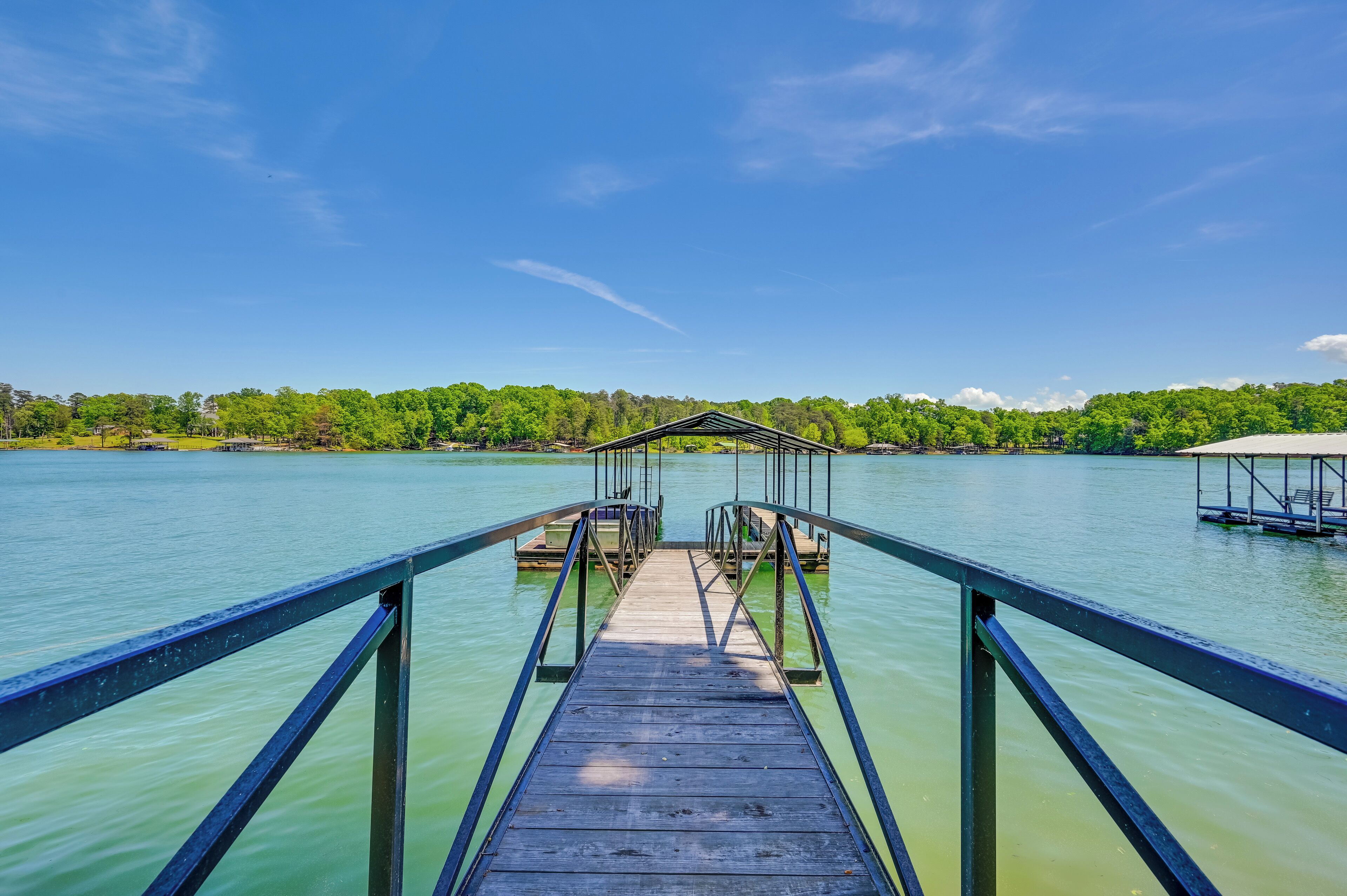 A wooden pier leading to a dock with a metal railing, surrounded by calm water and a forested shoreline.
