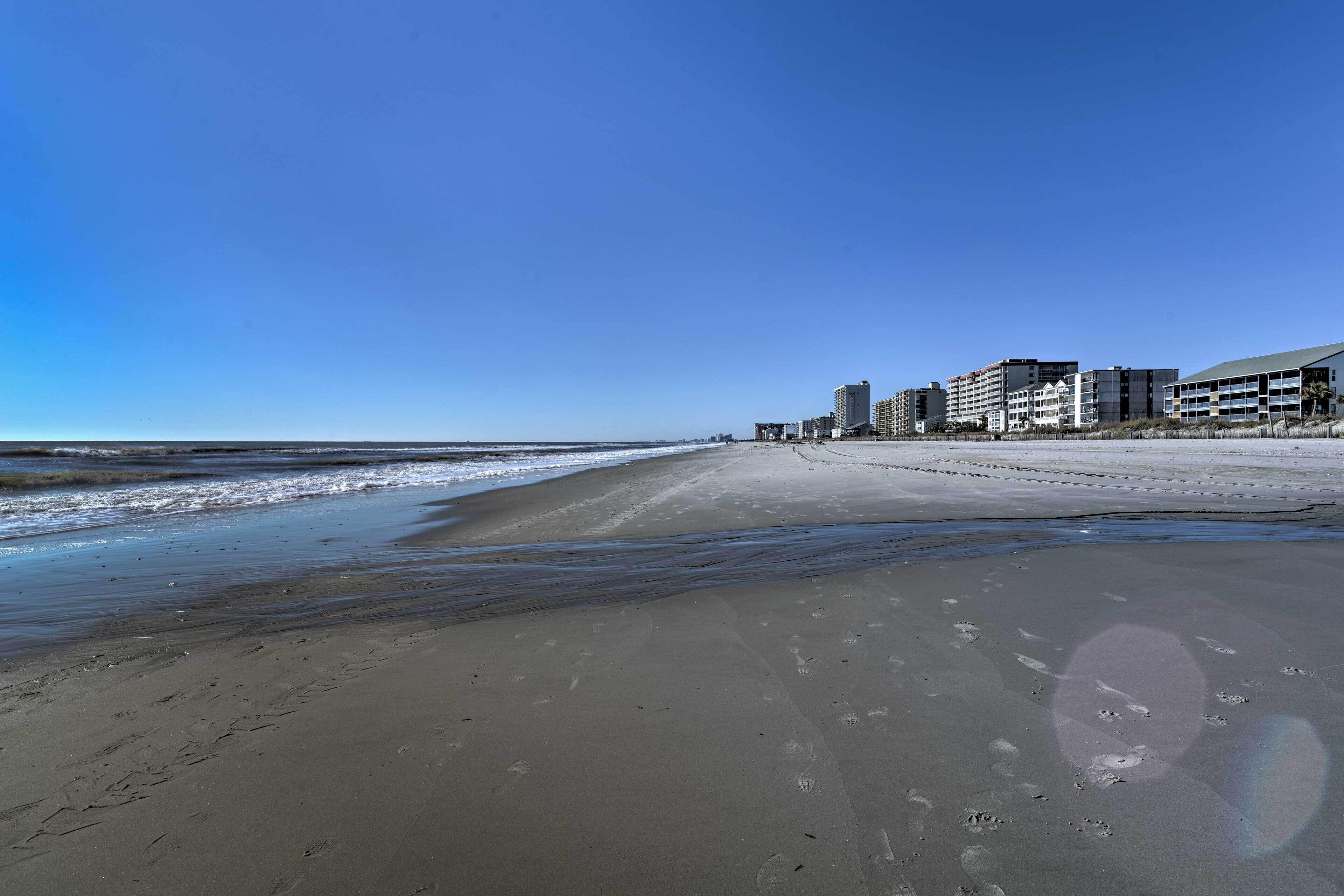 A beach with a clear sky, a sandy shore, and buildings in the background.