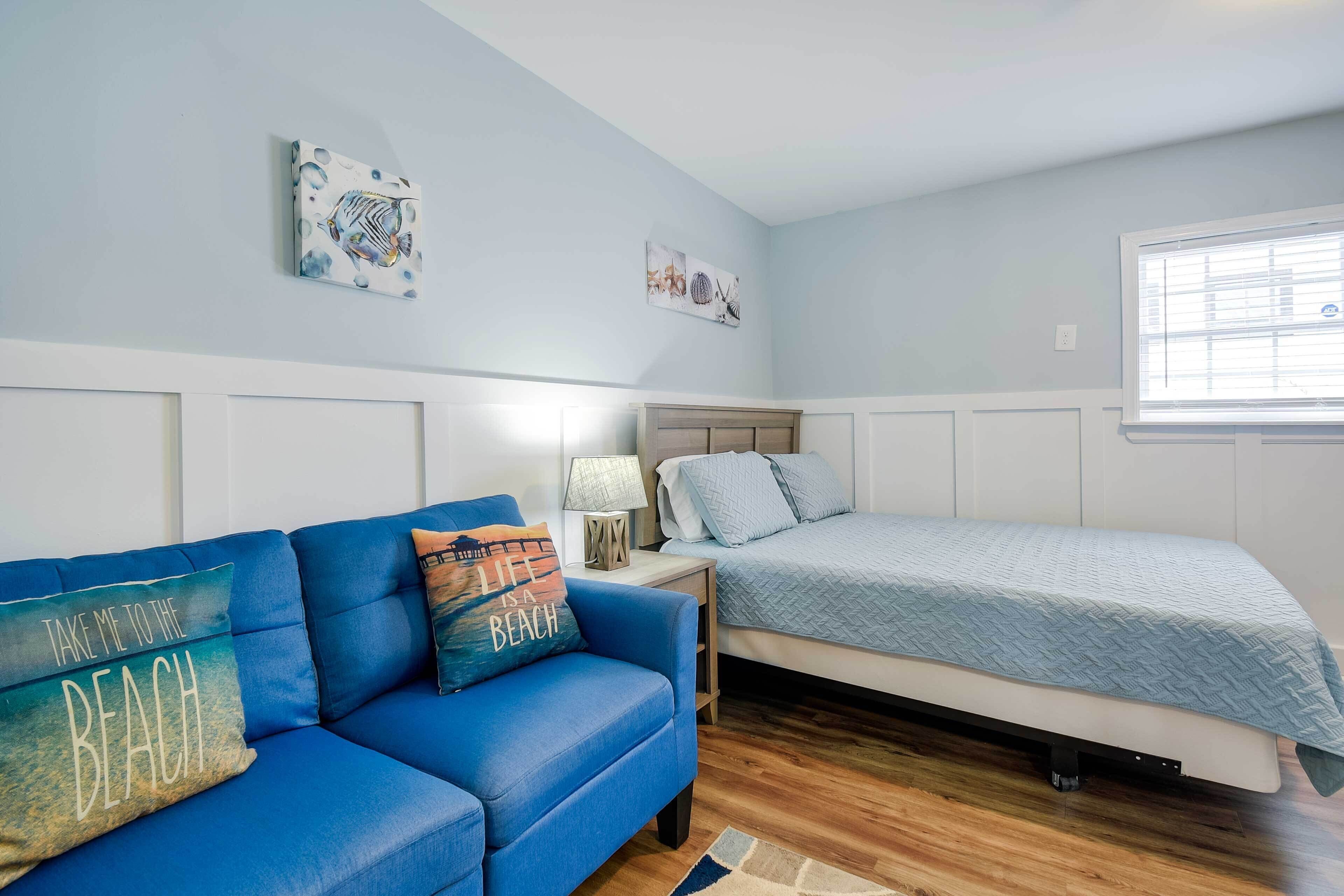 Ein Schlafzimmer mit einem blauen Sofa, einem Bett mit einer hellblauen Tagesdecke, einem Nachttisch mit einer Lampe und Wandbildern.