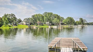 A lakefront property with a wooden dock extending into calm water, surrounded by lush greenery and a large house with multiple windows and a well-maintained lawn.