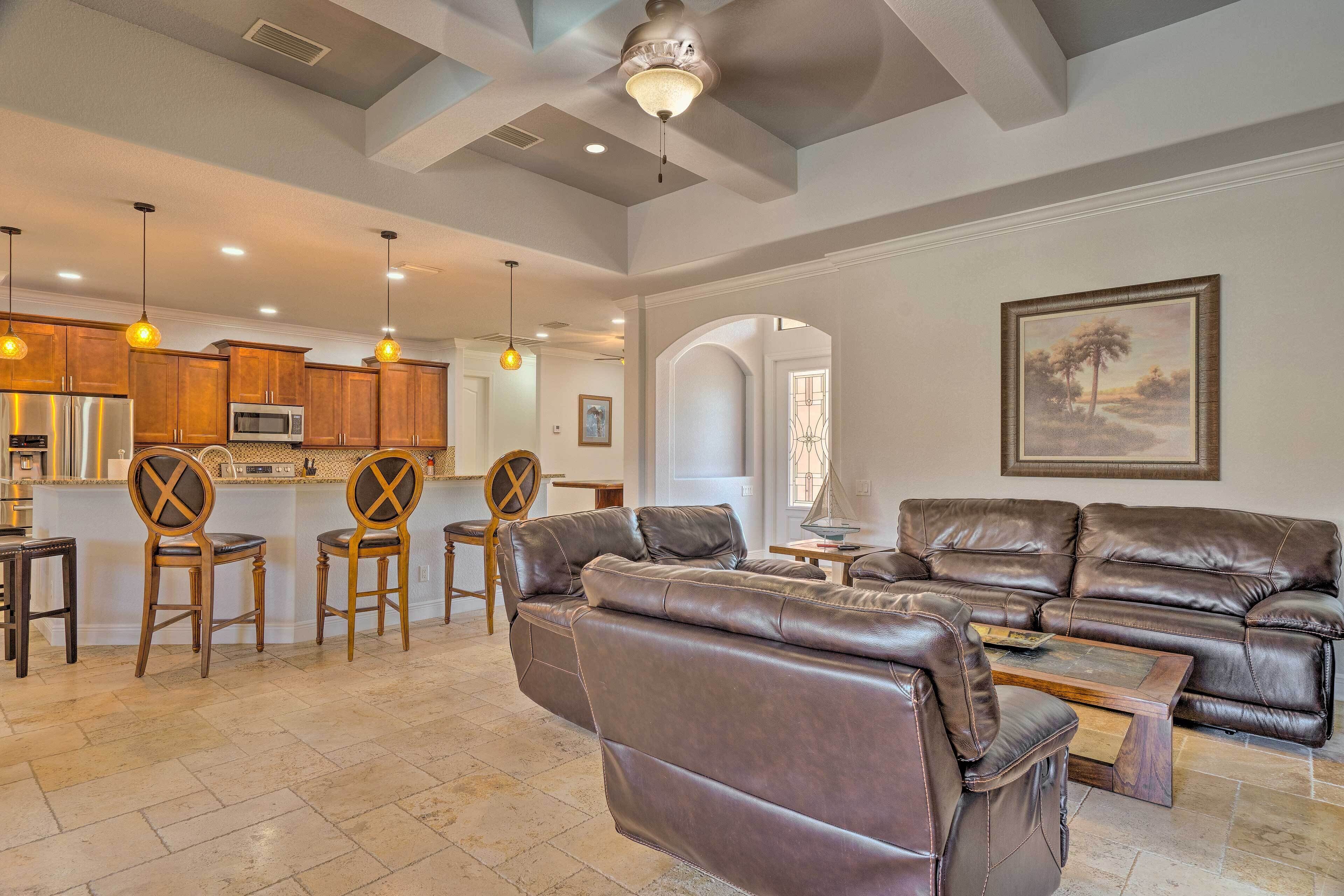 A spacious living area with a leather sofa, wooden chairs, a dining table, and a kitchen in the background.
