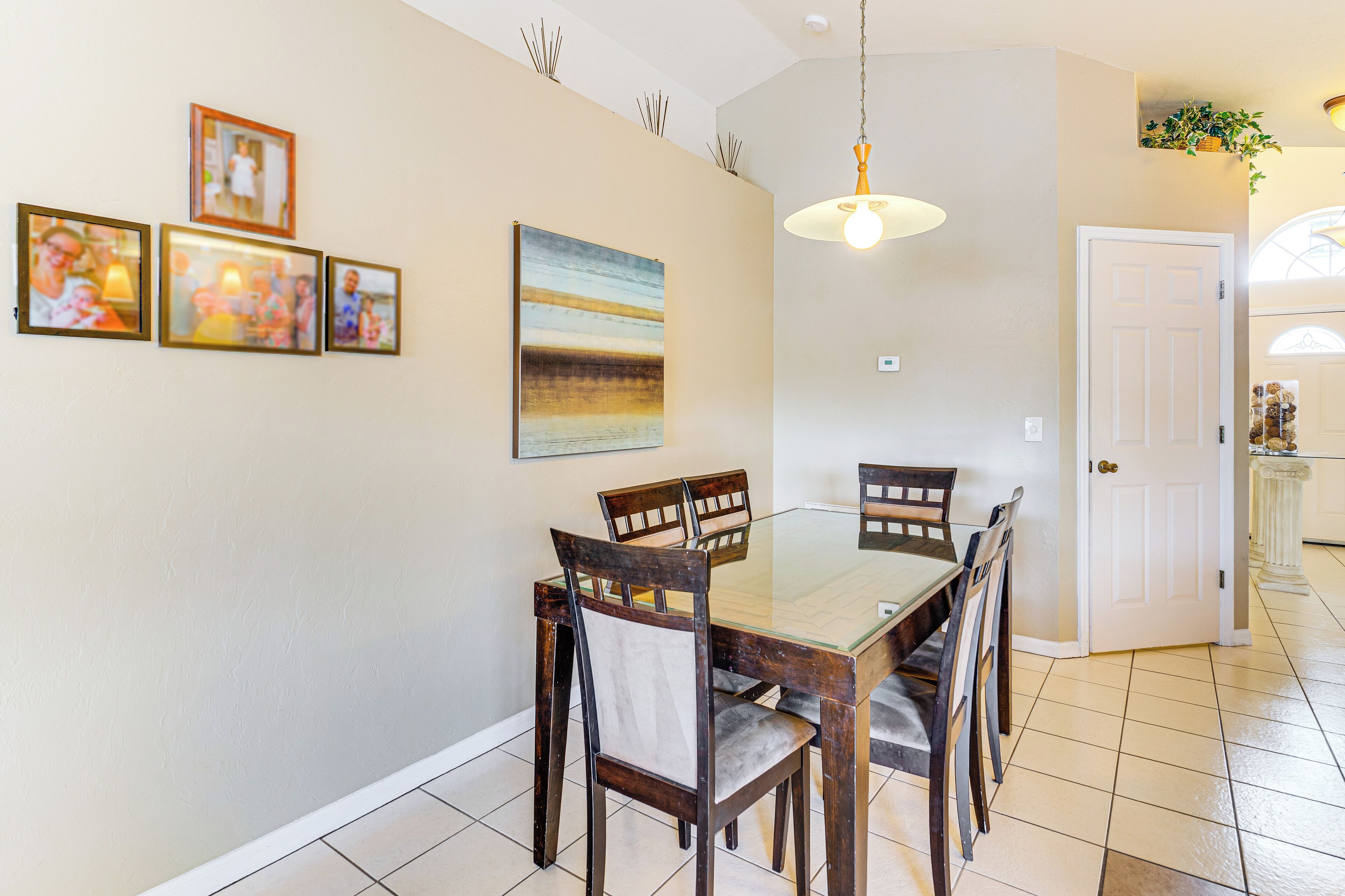 A dining area with a glass-top table, wooden chairs, and a pendant light. There are framed pictures on the wall and a door leading to another room.
