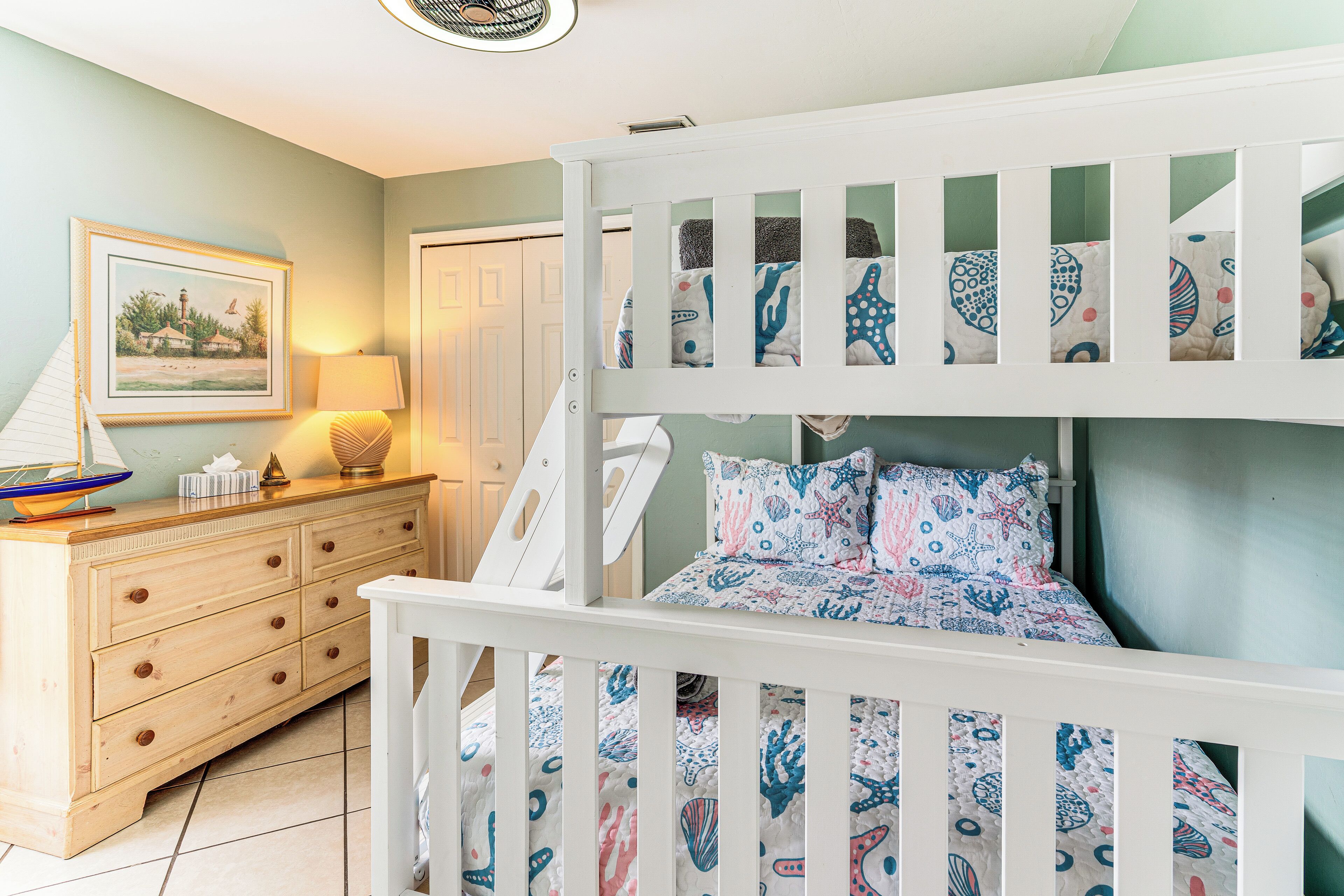 A bunk bed with a nautical theme, a dresser with drawers, a framed picture on the wall, and a lamp on the dresser.