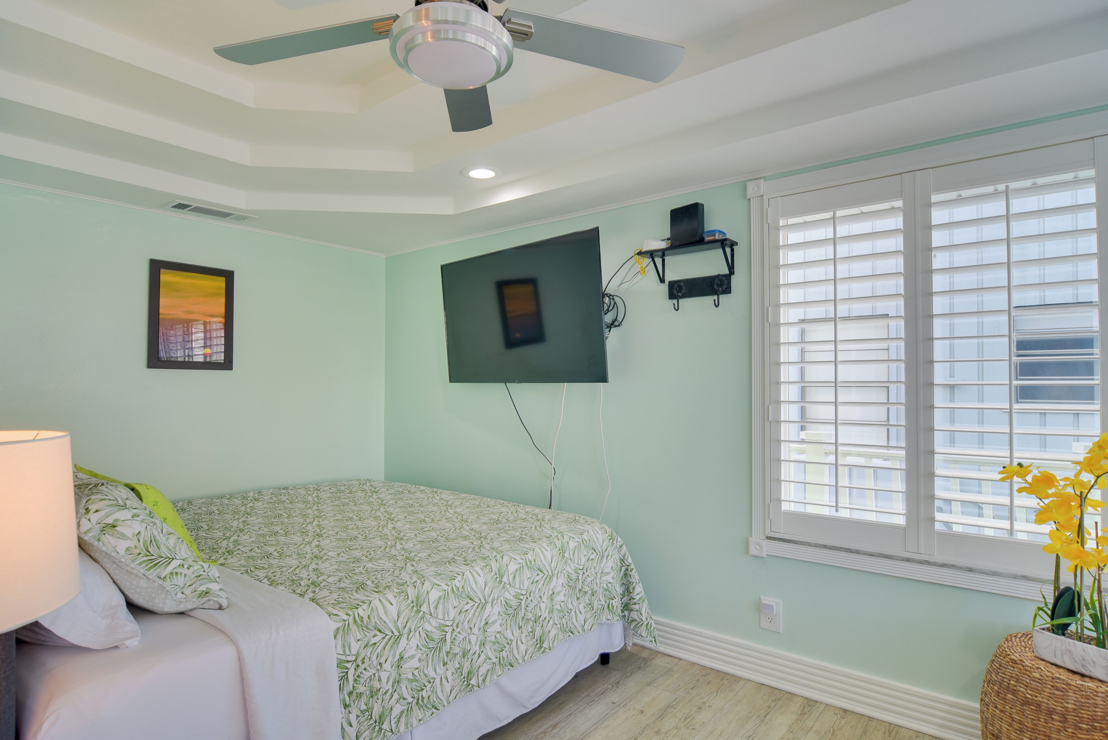 A bedroom with a bed, a ceiling fan, a window with blinds, and a wall-mounted shelf.