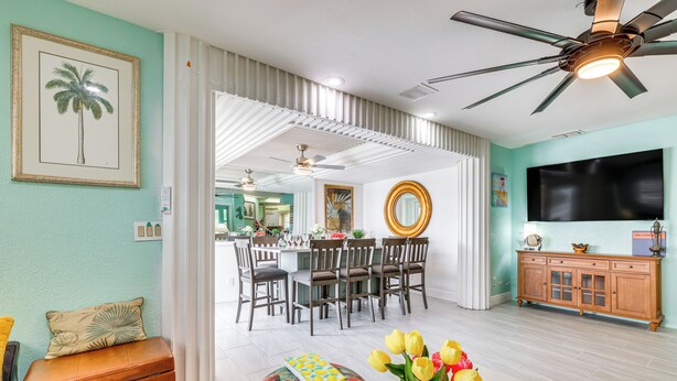 A dining area with a white ceiling fan, a wooden dining table with chairs, and a TV on the wall. - Breezy White Sands House Steps to Gulf Shore! (Clearwater Beach)