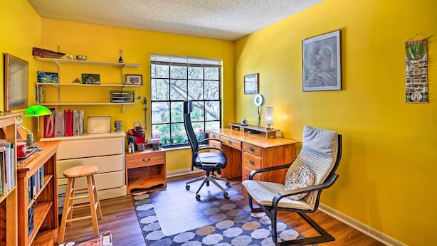A room with yellow walls, a wooden desk, a black office chair, a bookshelf, and a window with blinds. - Brandon Vacation Rental Home w/ Spacious Yard (Brandon)