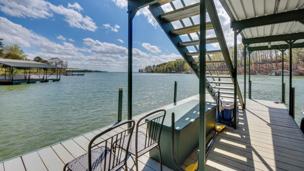 Eine überdachte Terrasse mit Metallgeländer, die auf einen ruhigen See, einen Steg und eine bewaldete Uferlinie blickt.