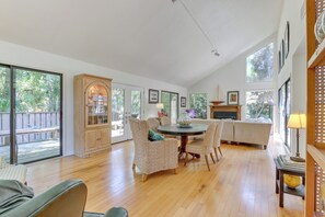 A spacious living area with a wooden dining table surrounded by wicker chairs, a sofa, and a fireplace. Large windows provide ample natural light and views of the outdoors. - 2 Mi to Beach: Hilton Head Home w/ Furnished Deck (Hilton Head Island)