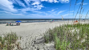 A beach with people under umbrellas, a lifeguard tower, and tall grass in the foreground. - Myrtle Beach Condo w/ On-site Pool & Beach Access (Myrtle Beach)