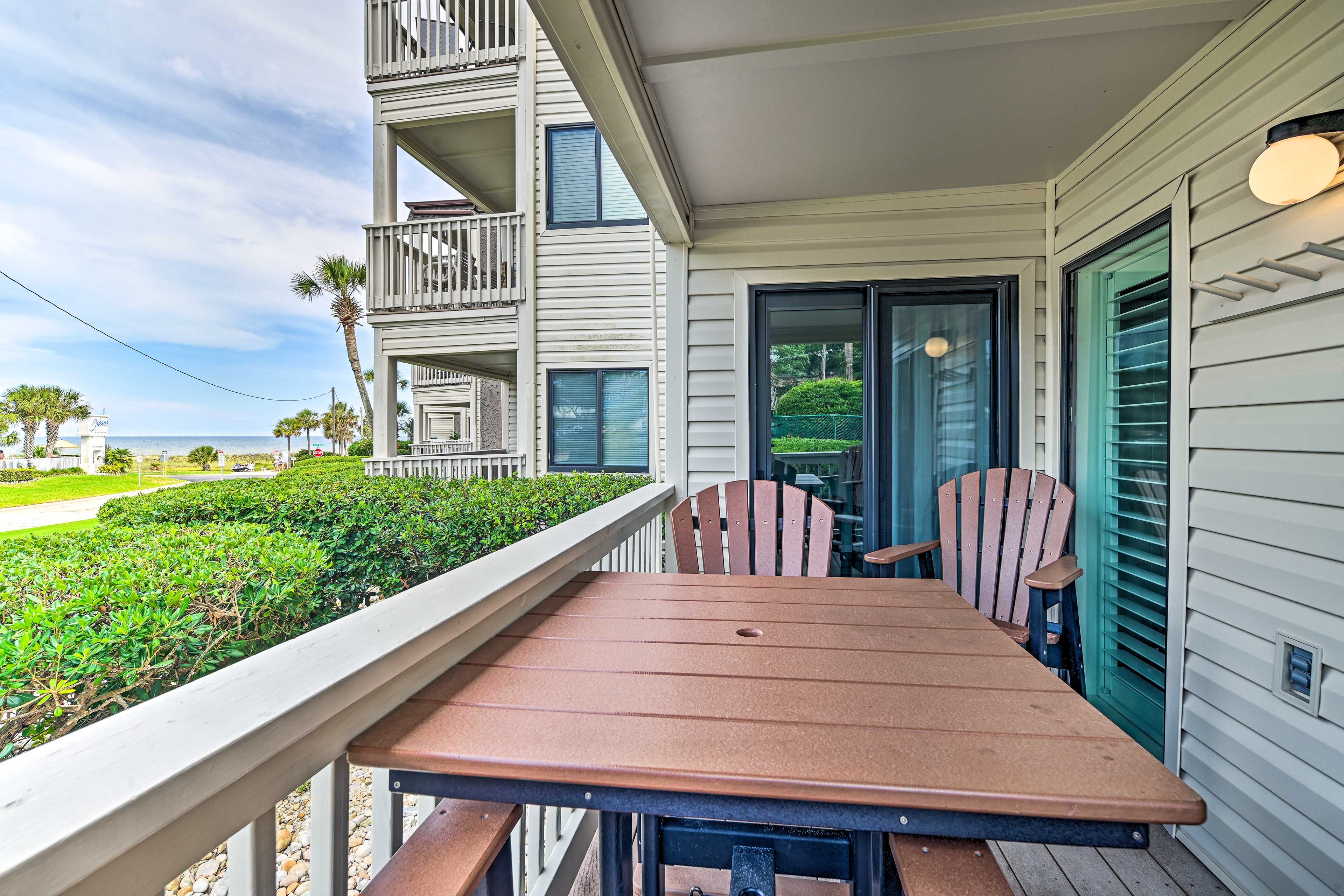 Une terrasse en bois avec une table et des chaises, donnant sur un espace aménagé avec vue sur un bâtiment et l’océan.