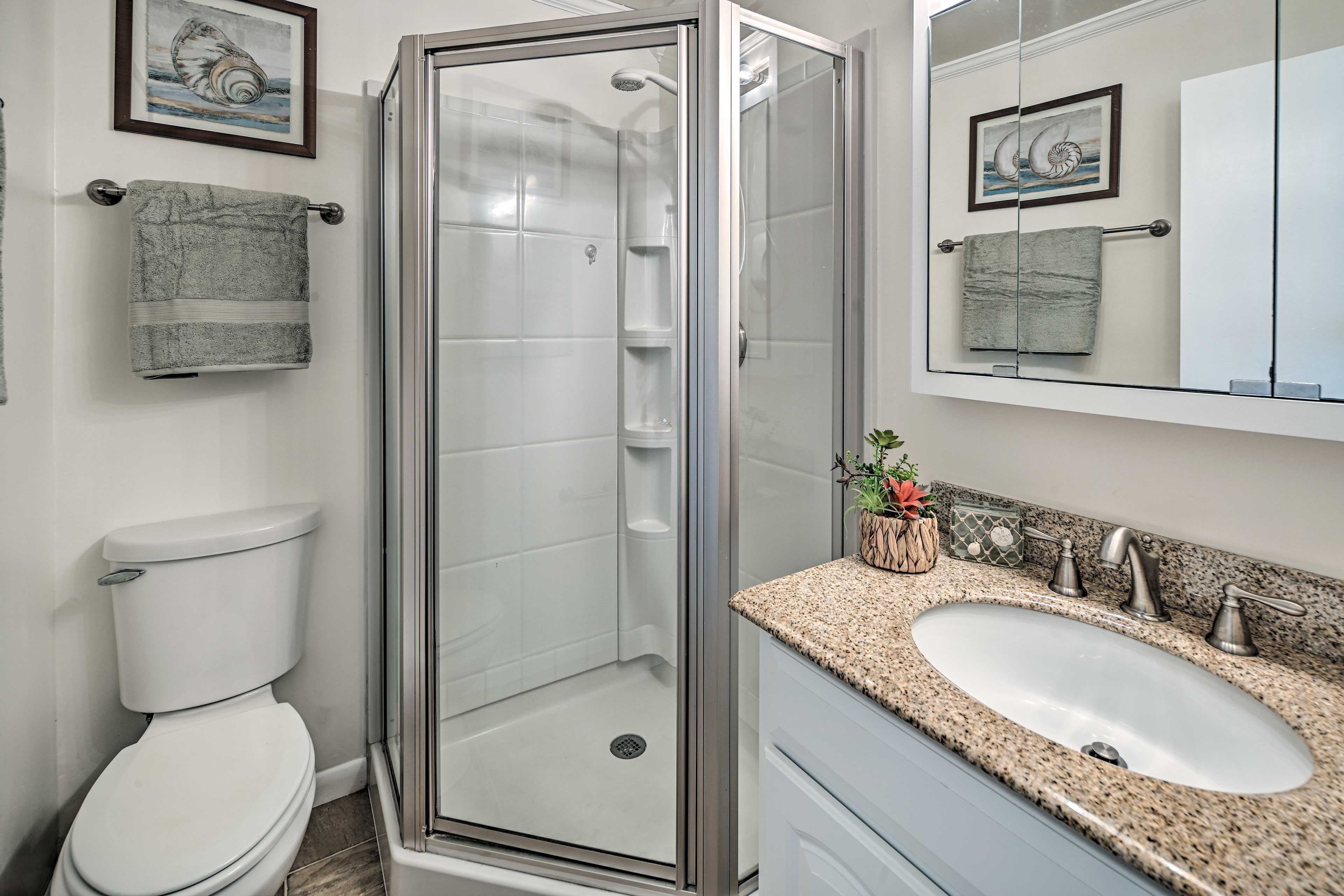 A bathroom with a glass-enclosed shower, a white toilet, a sink with a granite countertop, and two framed pictures on the wall.