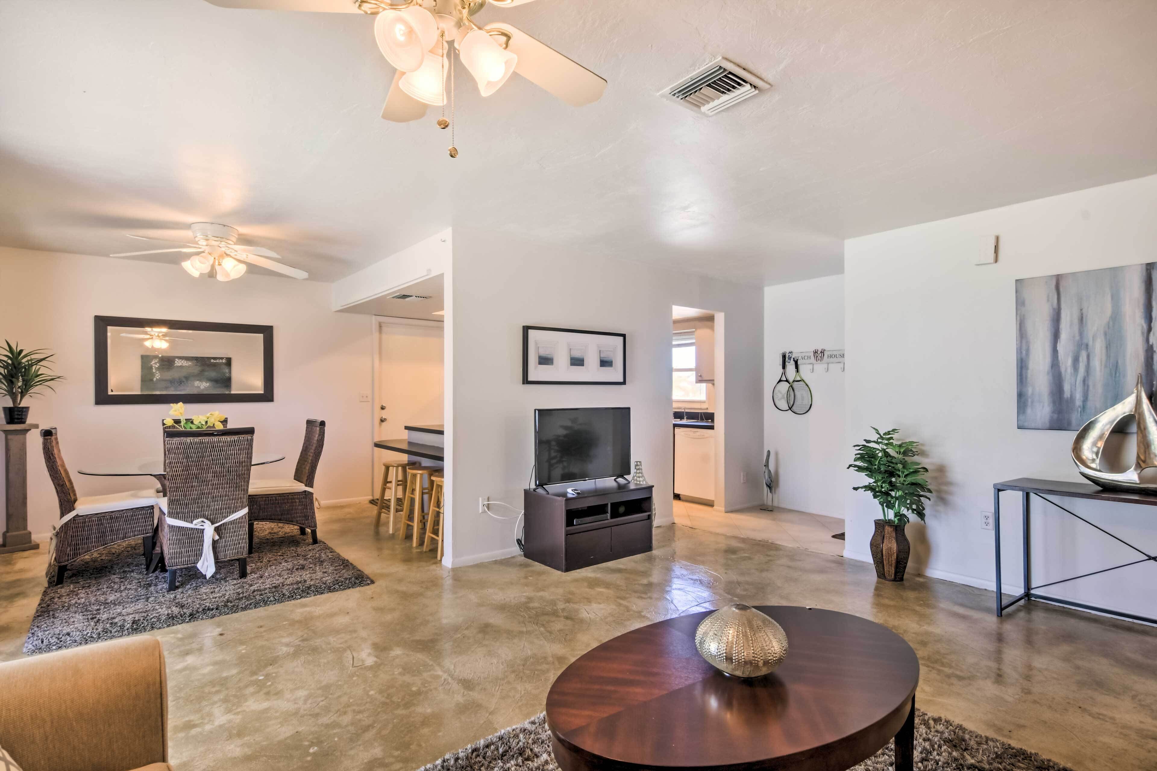 A spacious living room with a ceiling fan, a dining area with wicker chairs, a TV on a black stand, and a large abstract painting on the wall.