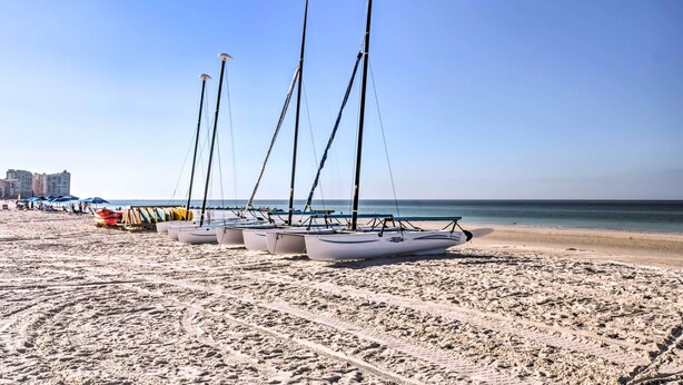 Ein Strand mit am Sand liegenden Segelbooten, einem klaren Himmel und Gebäuden im Hintergrund.