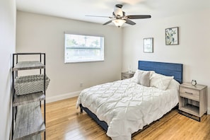 Quarto com cama, mesa de cabeceira, prateleira na parede, ventilador de teto e janela com persianas.