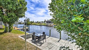 A patio with a table and chairs overlooking a body of water, surrounded by greenery and a clear sky.