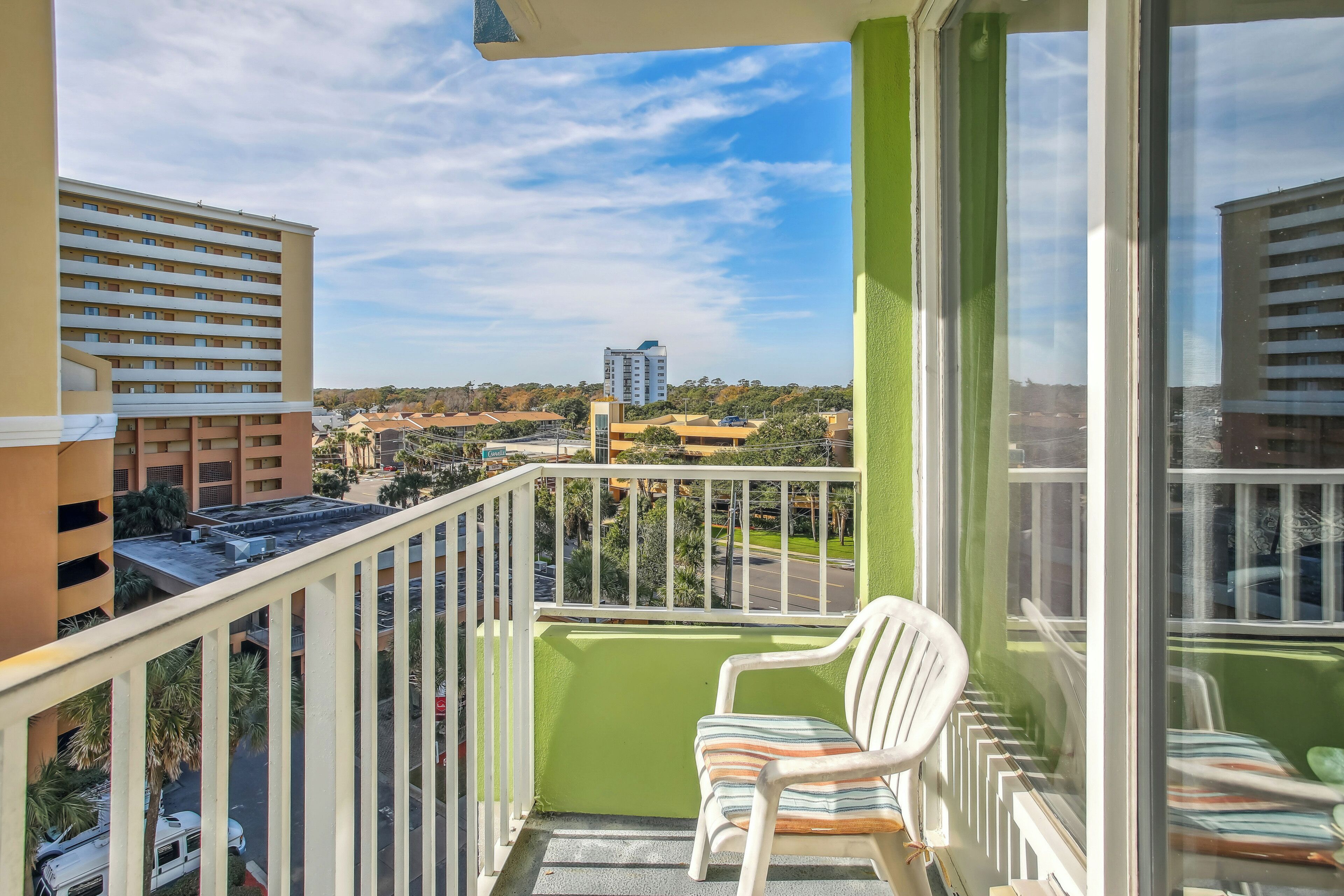 A balcony with a white chair, a green wall, and a view of surrounding buildings and a road.