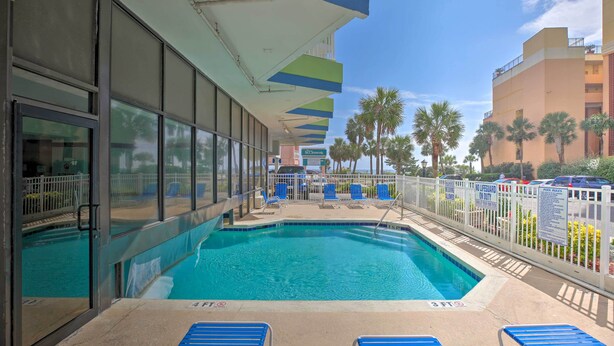 A swimming pool with blue chairs and umbrellas, surrounded by a white fence and palm trees. - Caravelle Tower Condo Rental: Walk to Beach (Myrtle Beach)