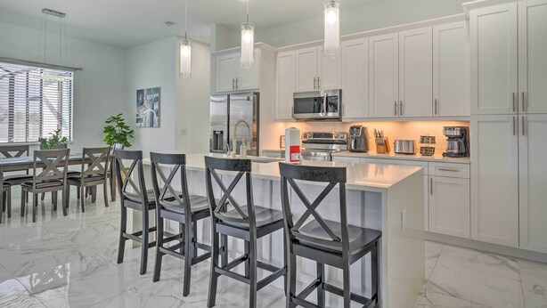 Una cocina moderna con armarios blancos, una isla central con taburetes de bar, electrodomésticos de acero inoxidable e iluminación colgante.