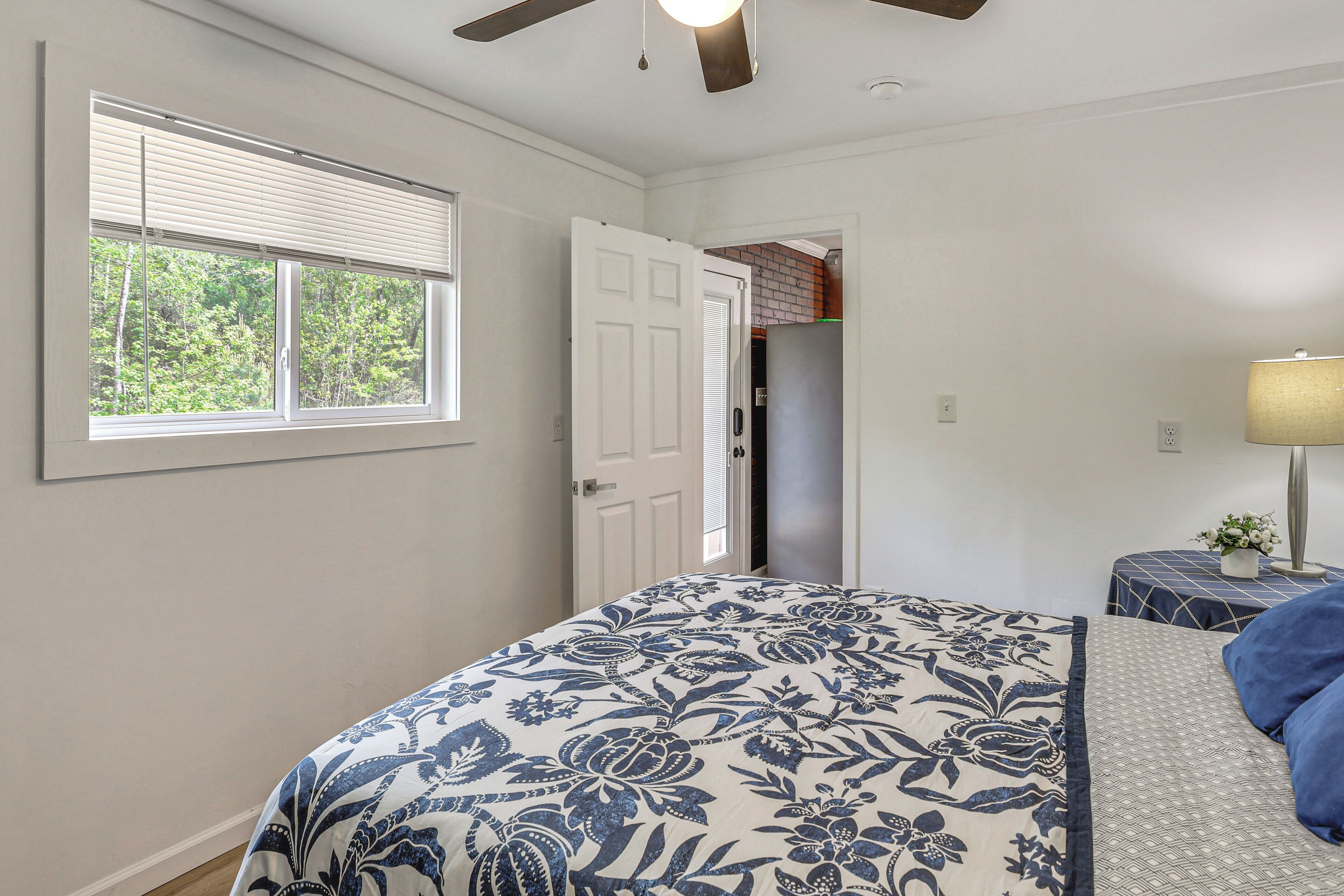 A bedroom with a bed, a window with blinds, a door, a ceiling fan, and a nightstand with a lamp.