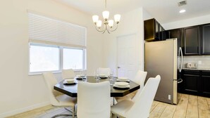 A dining area with a table set for four, white chairs, a chandelier, and a kitchen with dark cabinets and a stainless steel refrigerator. - 12 Mi to Disney: Home w/ Pool Access + Grill (Davenport)