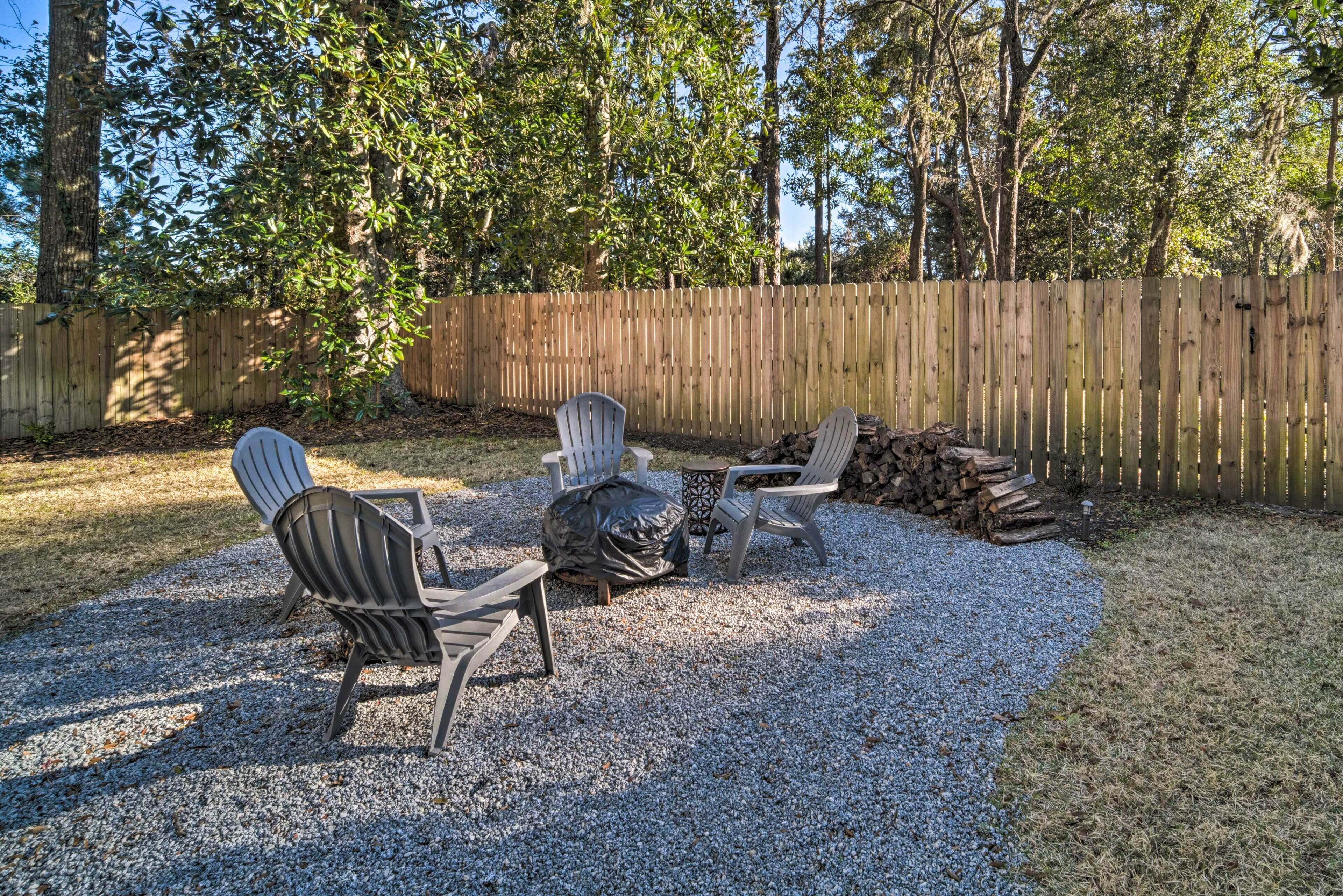Ein Garten mit Holzzaun, zwei Adirondack-Stühlen, einem kleinen Tisch und einem Feuerkorb mit Holzscheiten.