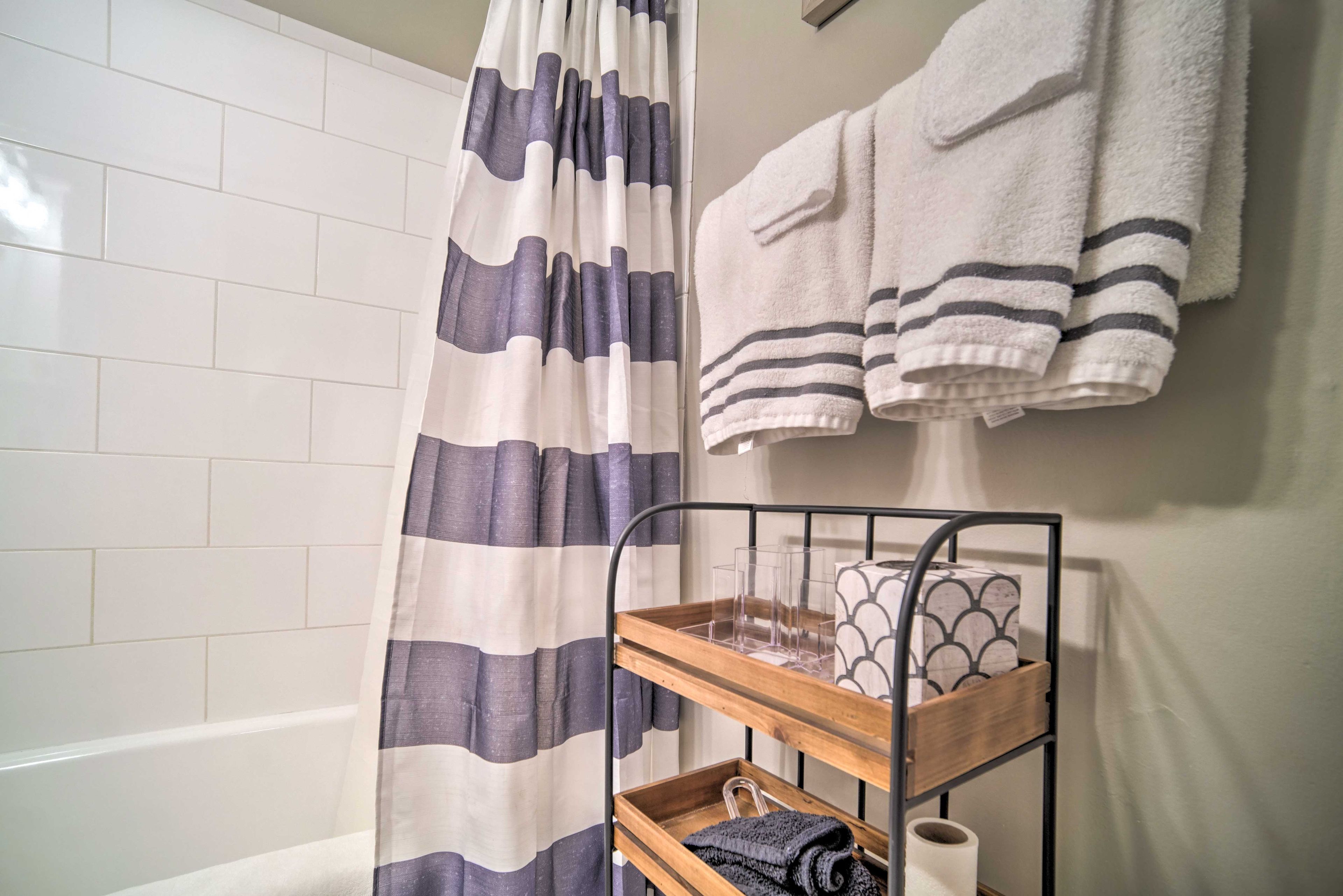 Une salle de bain avec un rideau de douche à rayures, un mur recouvert de carreaux blancs, un porte-serviettes avec deux serviettes et une étagère en bois où sont posés des articles de toilette.