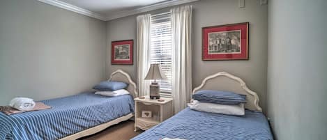 Een kamer met twee éénpersoonsbedden, een plafondventilator, een raam met gordijnen en twee ingelijste foto's aan de muur.