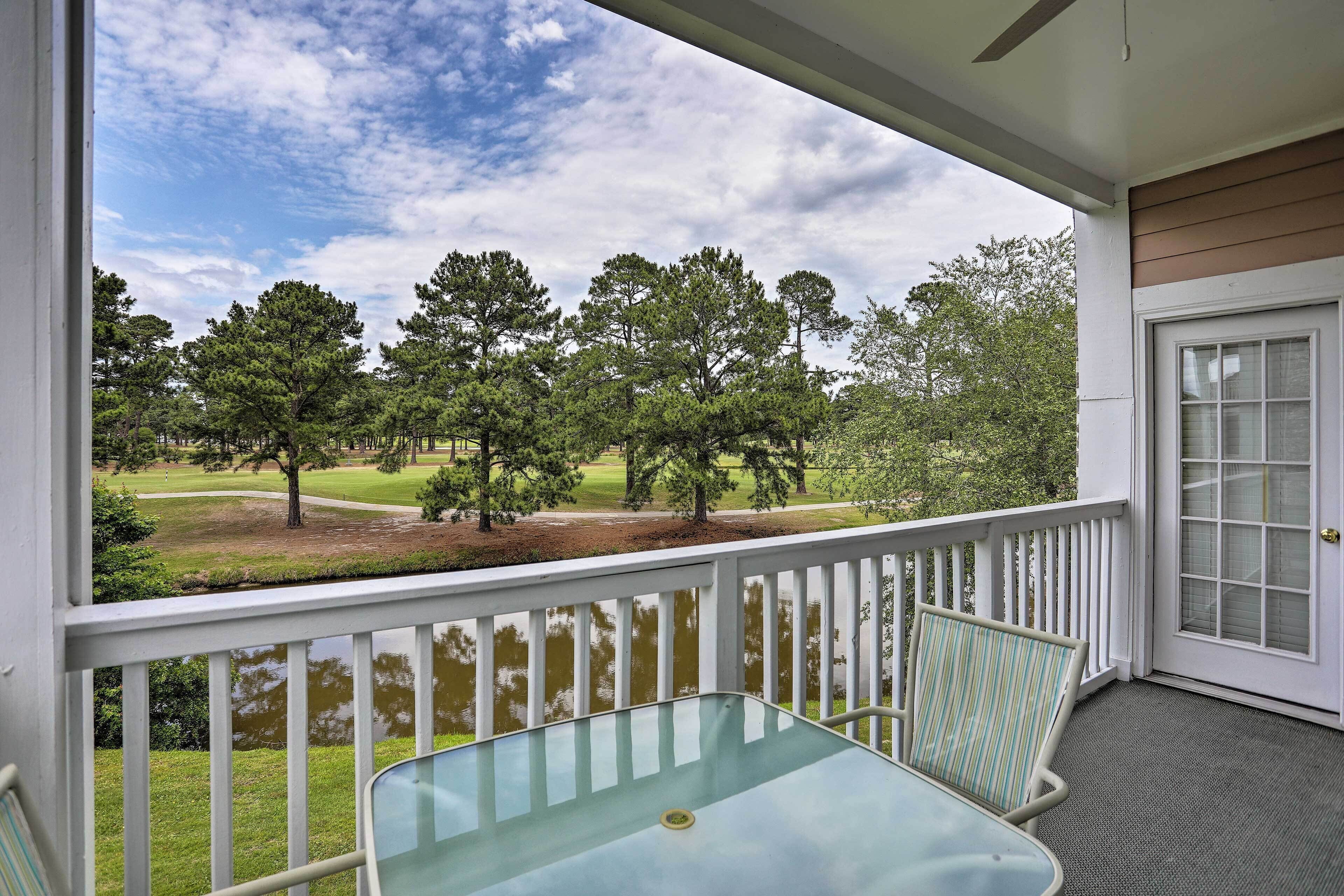 Une véranda avec une table et des chaises en verre, une balustrade blanche et une vue sur un terrain de golf et des arbres.