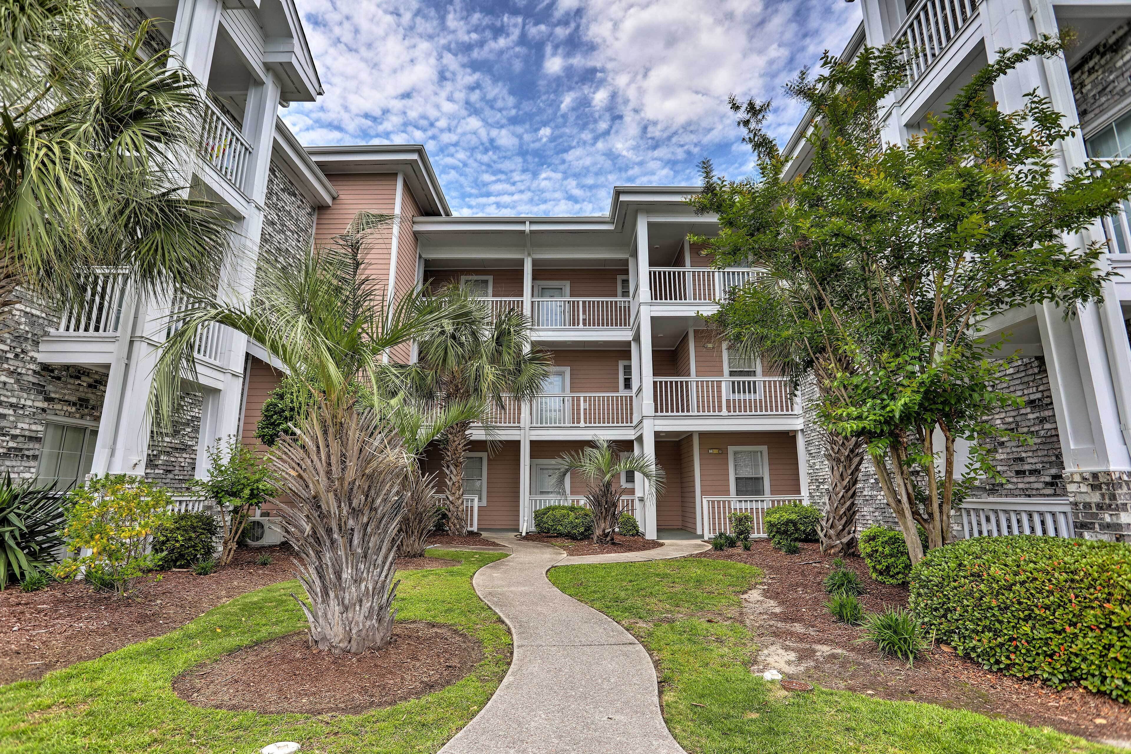 A multi-story residential building with balconies, a winding pathway, and various shrubs and trees.