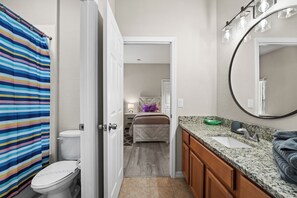 Une salle de bain avec un rideau de douche à rayures, des toilettes, un lit avec un oreiller violet, un lavabo avec un plan de travail en granit et des meubles en bois.
