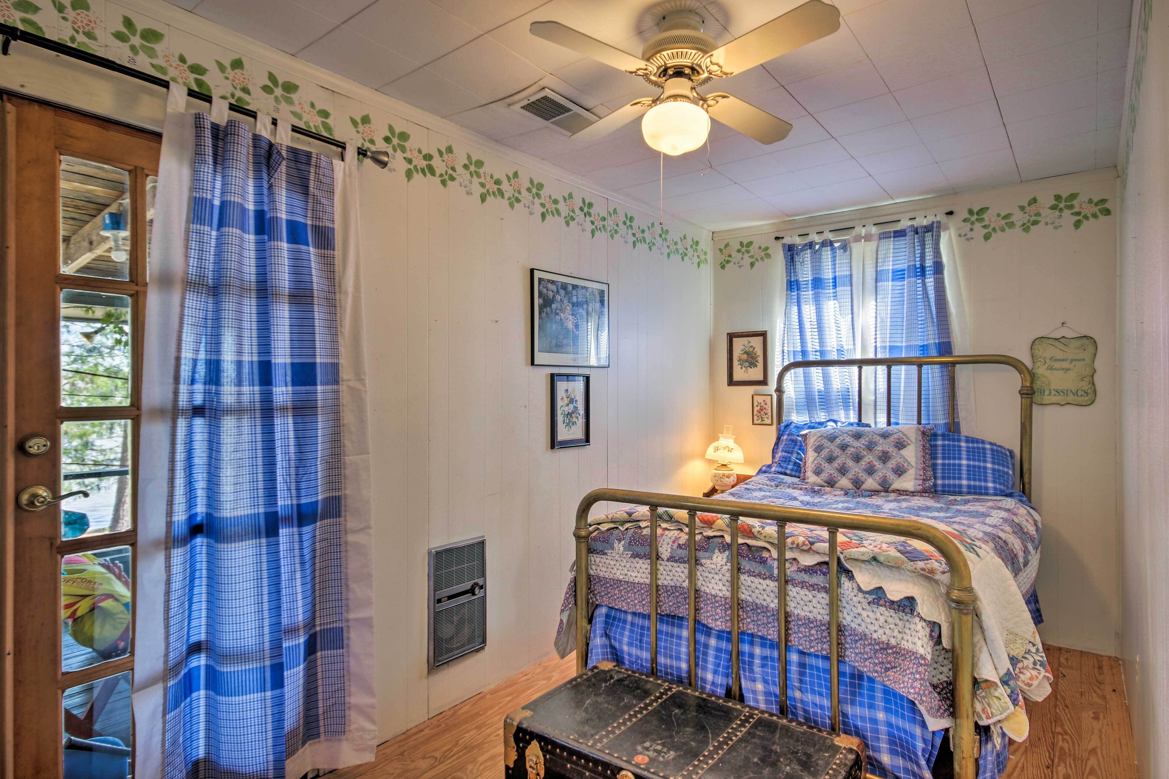 A bedroom with a bed, a nightstand, a window with curtains, a ceiling fan, and a door with a view of the outside.