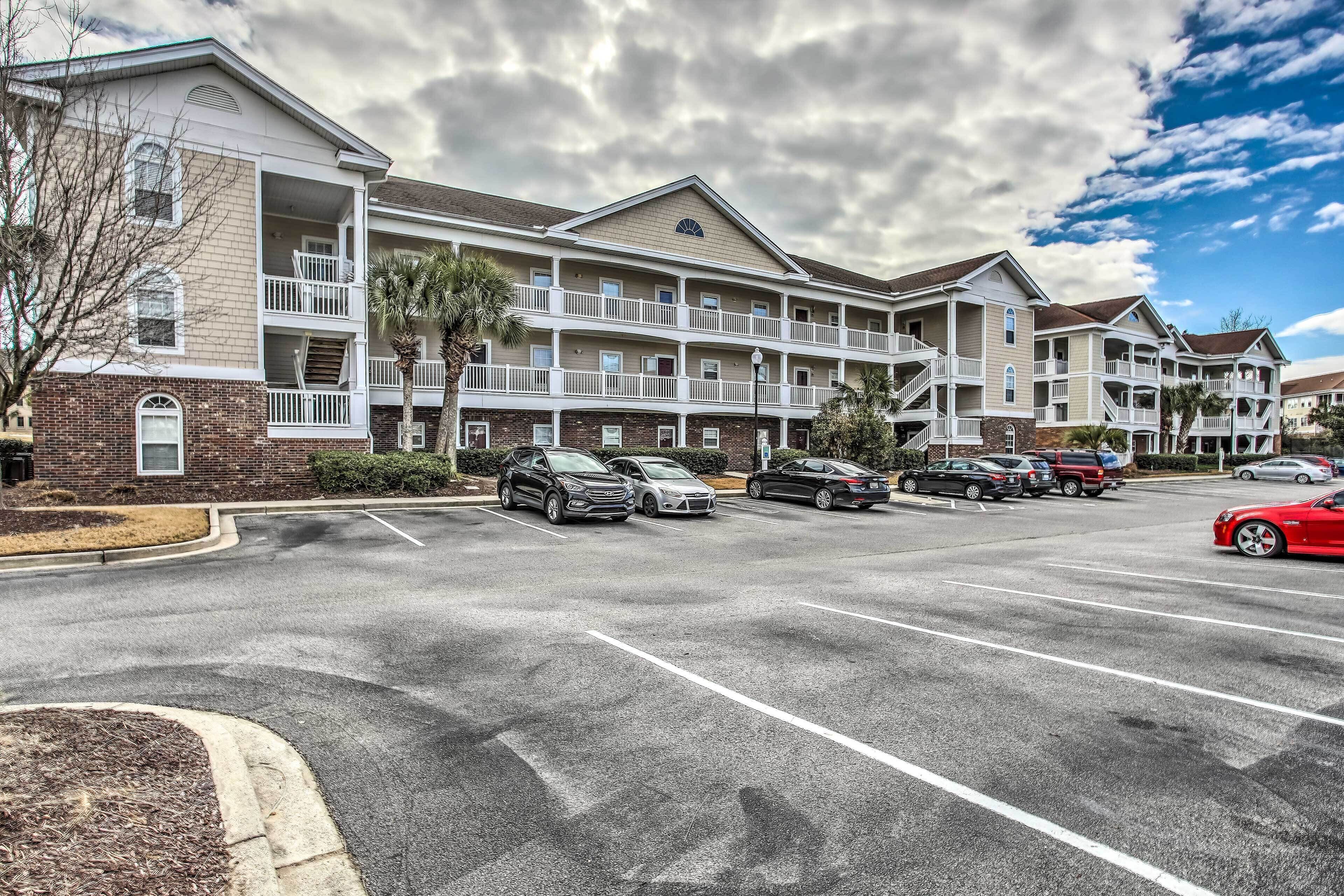 A multi-story hotel with a brick facade, multiple balconies, and a parking lot with several cars.