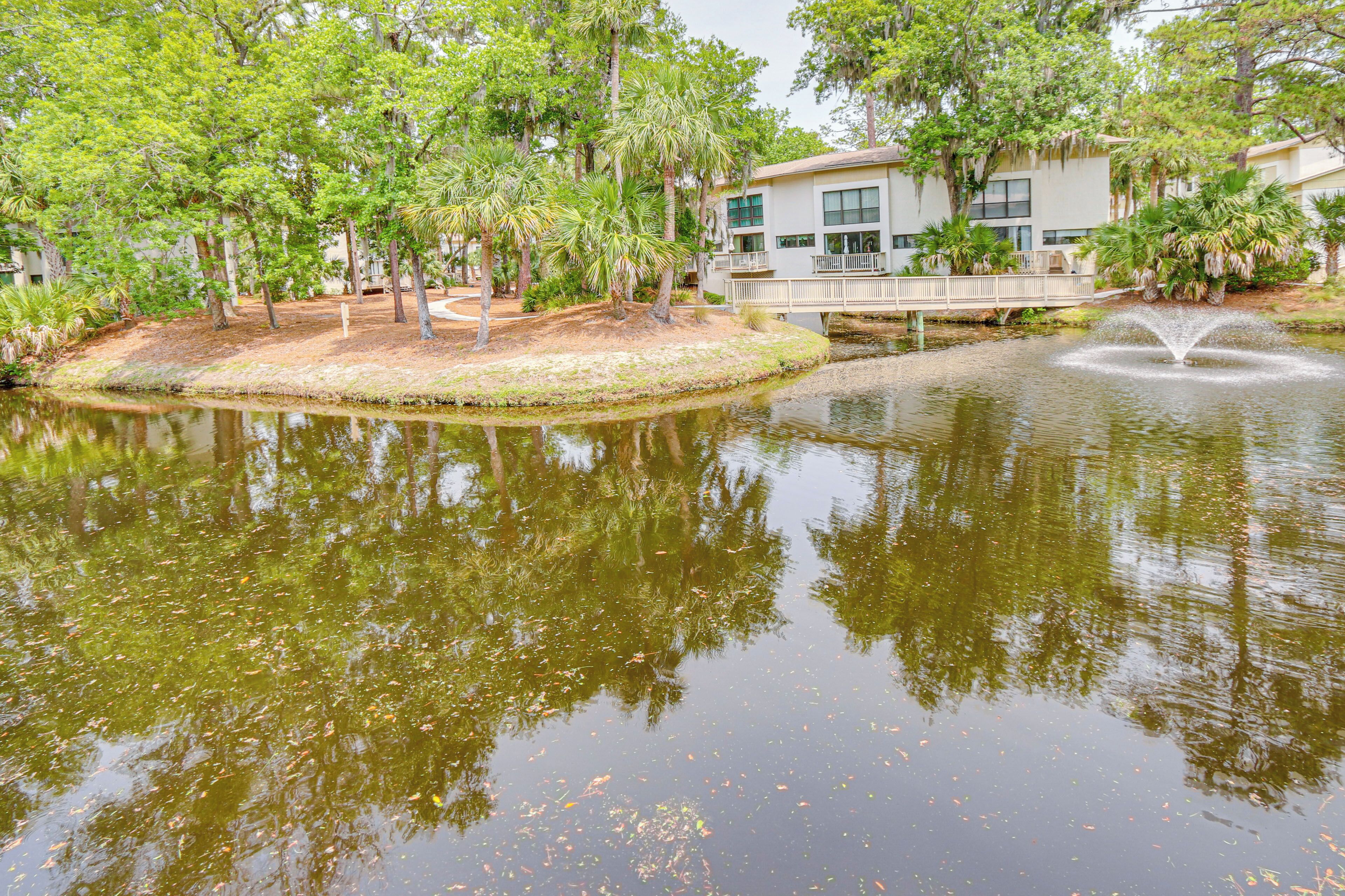 A serene pond with a fountain, surrounded by lush greenery and a bridge.