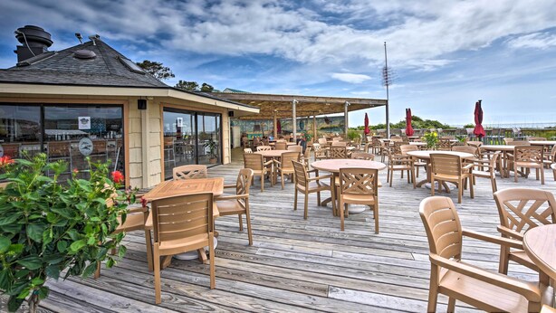 An outdoor patio area with wooden tables and chairs, a covered seating area, and a view of the sea. - Pool, Spa & Dining: Beach Condo in Hilton Head! (Hilton Head Island)