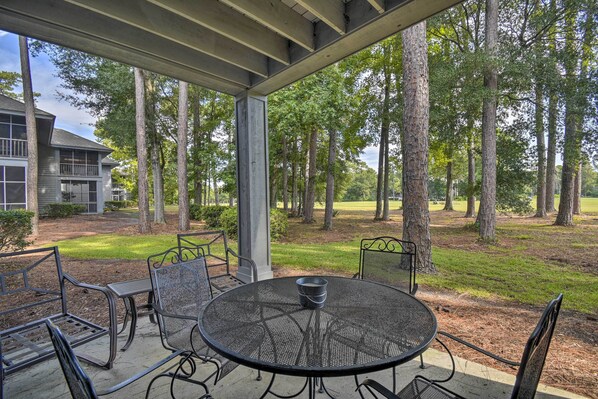 An outdoor patio with a round table and metal chairs, surrounded by trees and a building in the background. - 4 Mi to Ocean: Golf Resort Condo in N Myrtle Beach (North Myrtle Beach)