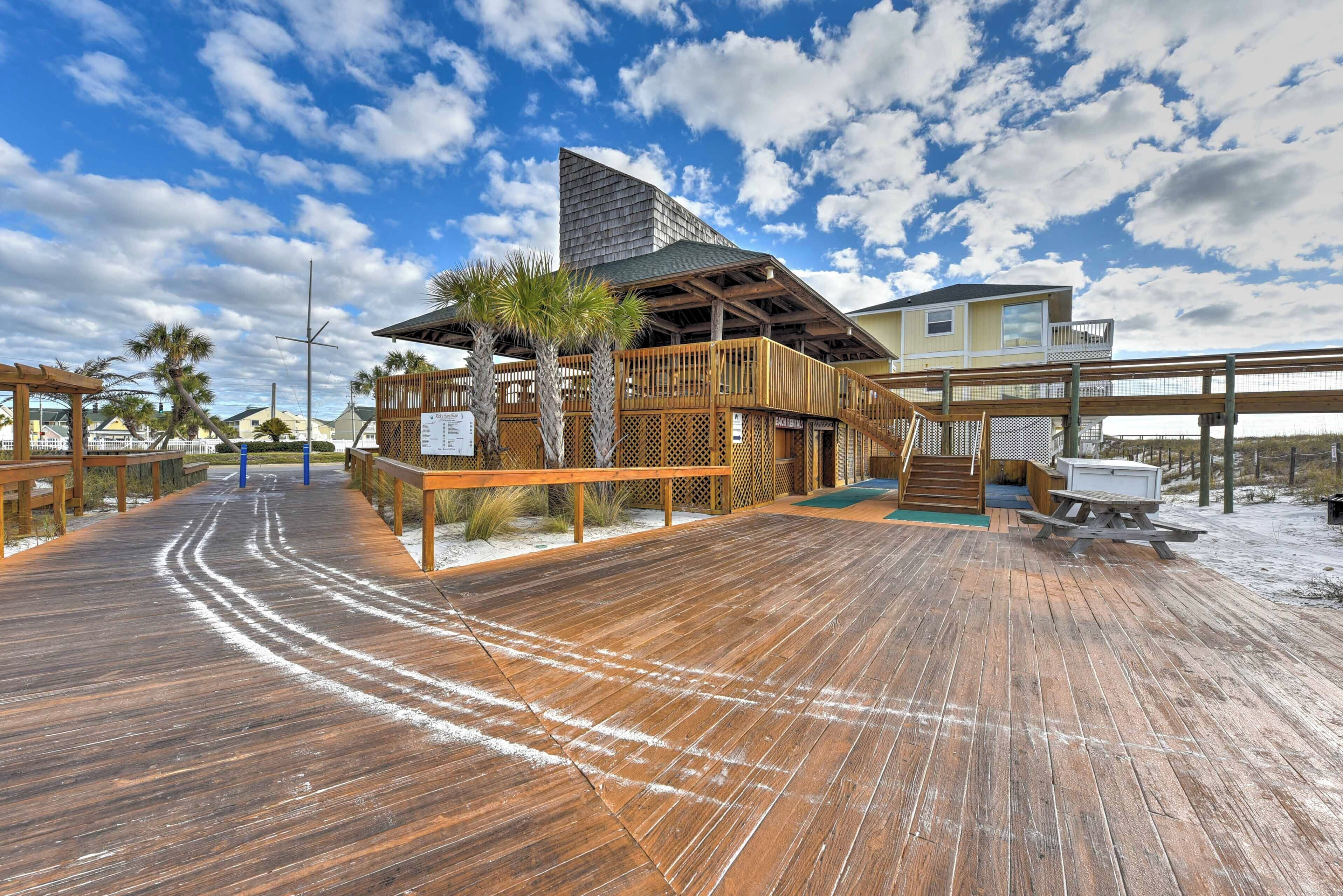 A wooden boardwalk leading to a beach house with a balcony, palm trees, and a clear sky.