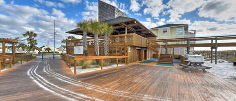 A wooden boardwalk leading to a beach house with a balcony, palm trees, and a clear sky.