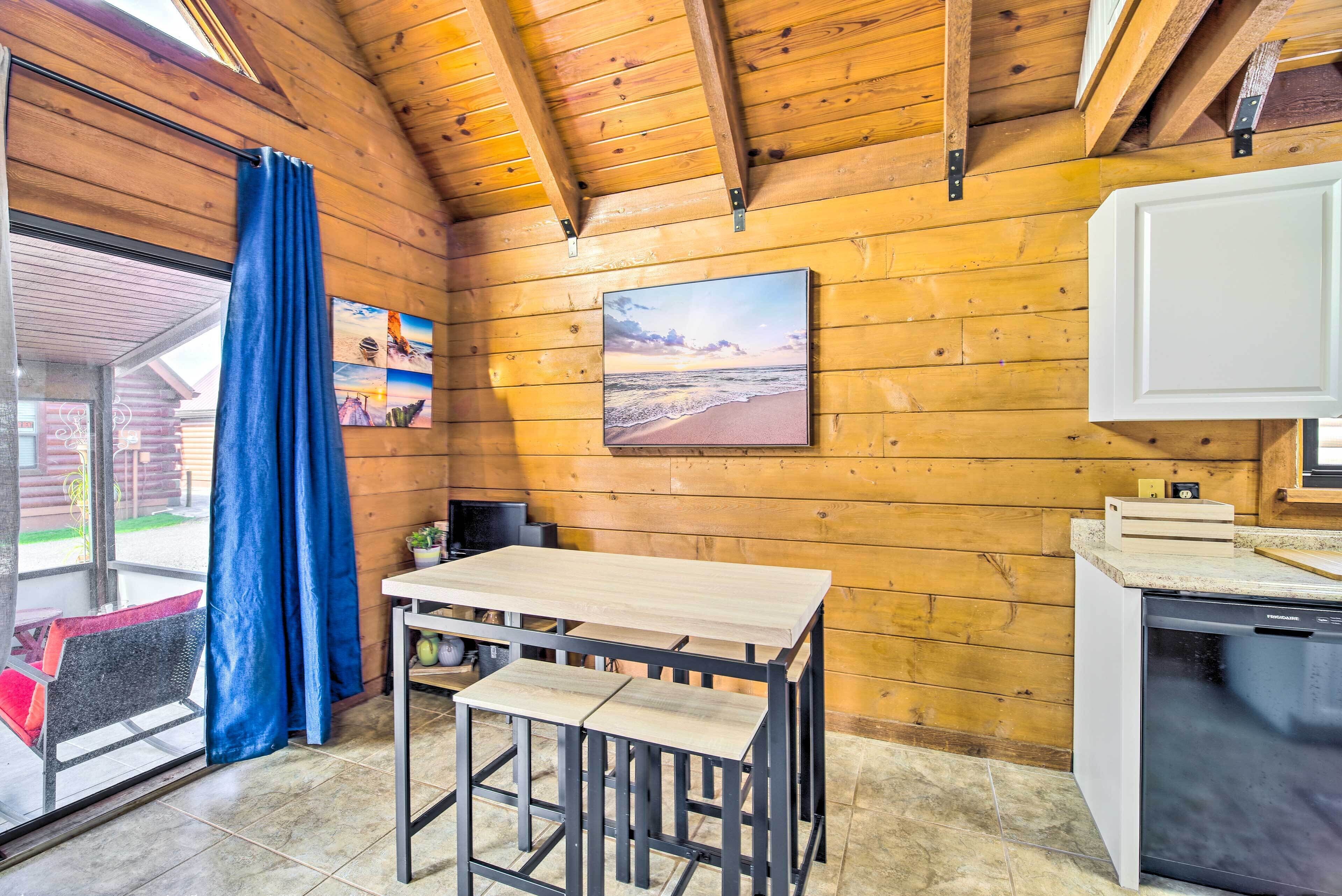 L’intérieur d’un chalet en bois comprend une table à manger, des chaises, un réfrigérateur et une fenêtre ornée d’un rideau bleu.