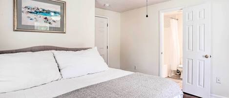 Un dormitorio con cama, ventilador de techo, un cuadro enmarcado en la pared y una puerta que da a un baño.