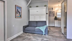 A modern living room with a gray sofa, a white built-in cabinet with plants, and a kitchen in the background. - Cozy Jacksonville Studio - 10 Miles to Downtown! (Jacksonville)