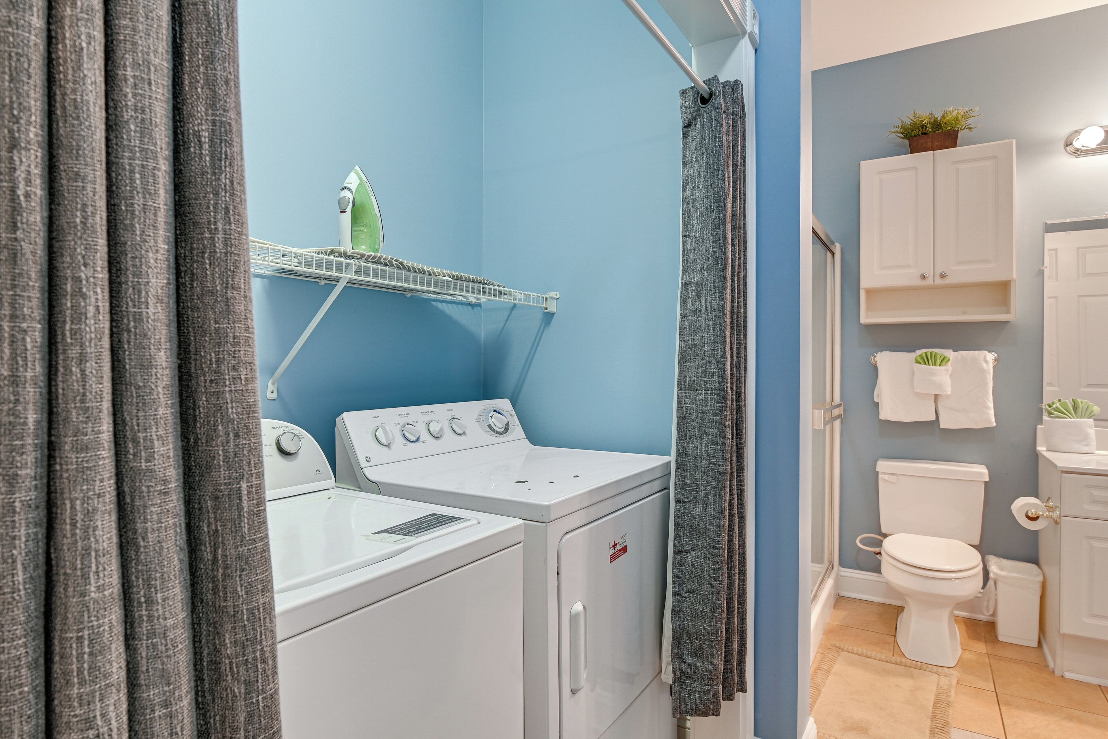 A laundry area with a washing machine and dryer, a shelf with a plant, and a toilet with a towel rack.