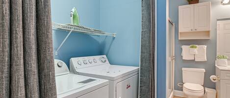 A laundry area with a washing machine and dryer, a shelf with a plant, and a toilet with a towel rack.