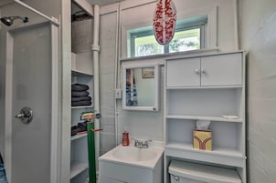 Un baño con ducha, lavamanos y espejo. Hay un perchero con toallas y una caja de pañuelos en los estantes.