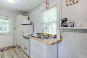 Une cuisine de taille réduite avec des meubles blancs, une cuisinière, un four à micro-ondes et un évier. On y trouve une fenêtre avec des stores, une étagère avec une machine à café et une boîte de café Maxwell House.
