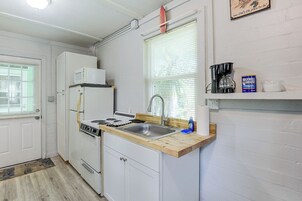 Cozinha compacta com armários brancos, fogão, micro-ondas e pia. Há uma janela com persianas, uma prateleira com cafeteira e uma lata de café Maxwell House.