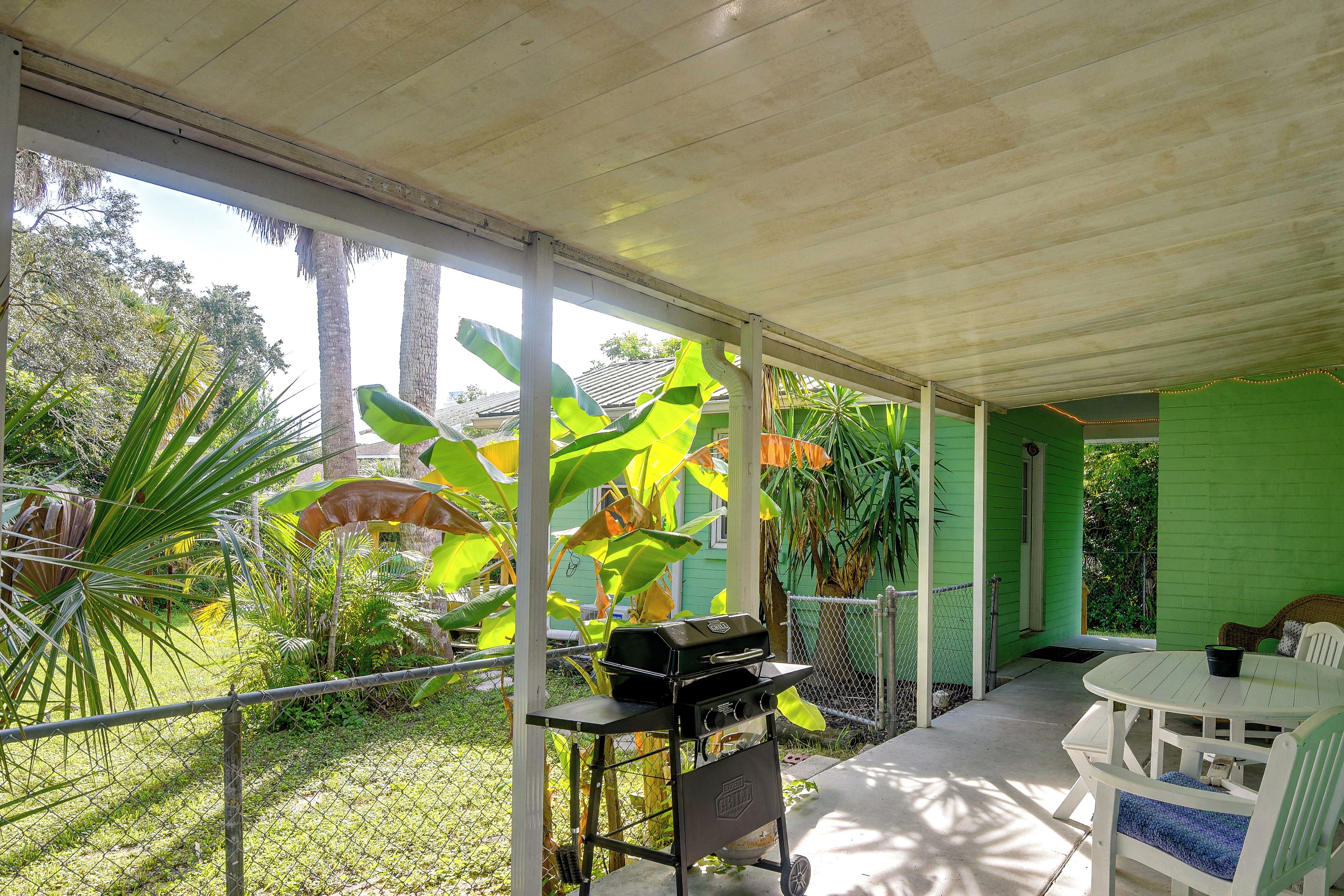 A covered outdoor area with a grill, a table, and chairs, surrounded by lush greenery and a green building.