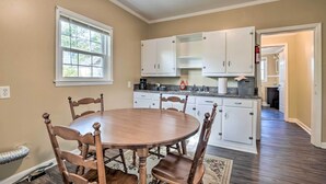A dining area with a round table and six chairs, a kitchen with white cabinets, and a window with blinds.